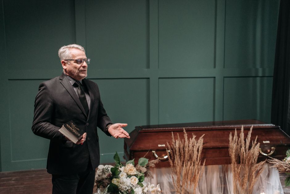 A man with grey hair and glasses dressed in formal black attire, including a suit and tie, stands in a dimly lit room with teal panelled walls. He is holding a small notebook in his left hand and gest