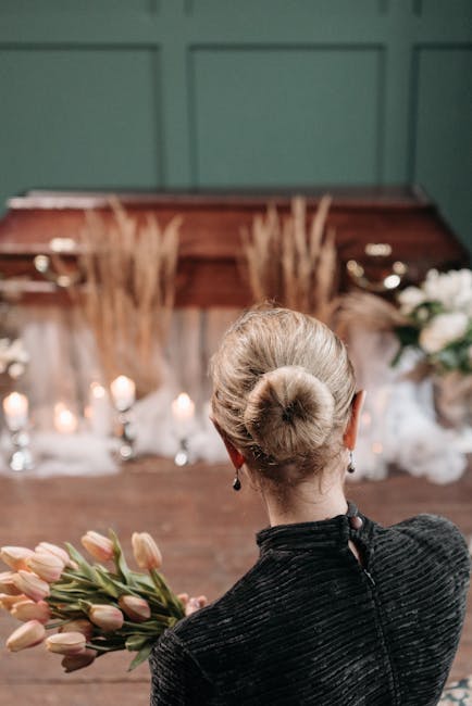A woman with blonde hair styled in a neat bun, dressed in a black textured jacket, holds a bouquet of pink tulips with green stems in her right hand. She is facing away from the camera, observing a fl