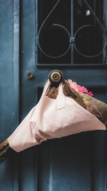 A person wearing brown gloves is using a brass door knocker to hold a wrapped floral gift delivery, featuring a bouquet of vibrant pink flowers inside brown paper, positioned against a dark blue paint