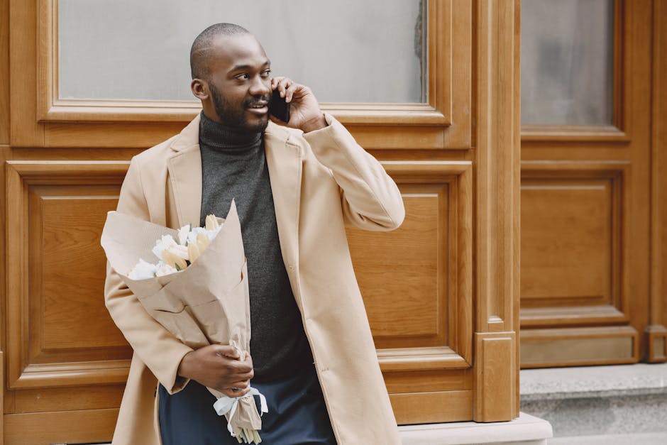 A man with dark skin, a beard, and a shaved head is standing outdoors against wooden paneling on a building. He is wearing a beige overcoat over a dark grey turtleneck sweater. He is holding a bouquet