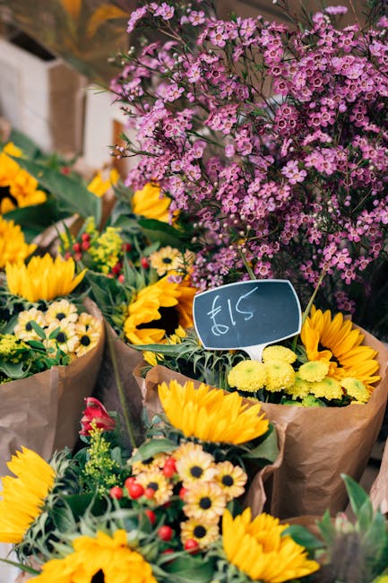 A vibrant display of fresh flower bouquets arranged on a market stand, featuring bright yellow sunflowers, small yellow and white daisies, and sprigs of greenery wrapped in brown paper. Behind the bou