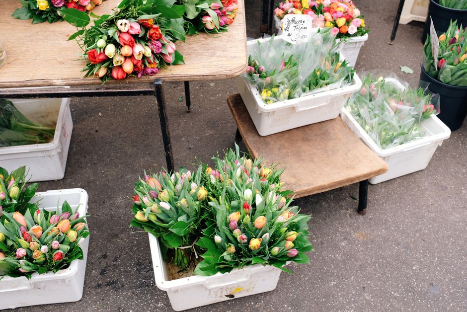 A display of fresh flower bouquets and tulips arranged in white plastic crates and on small wooden tables at an outdoor market stall. The bouquets feature a variety of tulips in pastel shades of pink,