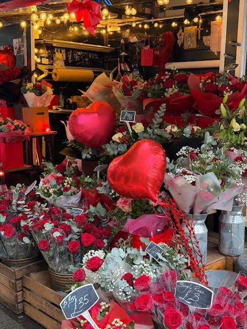 A vibrant display of fresh floral arrangements showcasing red and white roses, baby's breath, and greenery, arranged in baskets and wrapped in pink and white paper. Among the flowers are red heart-sha