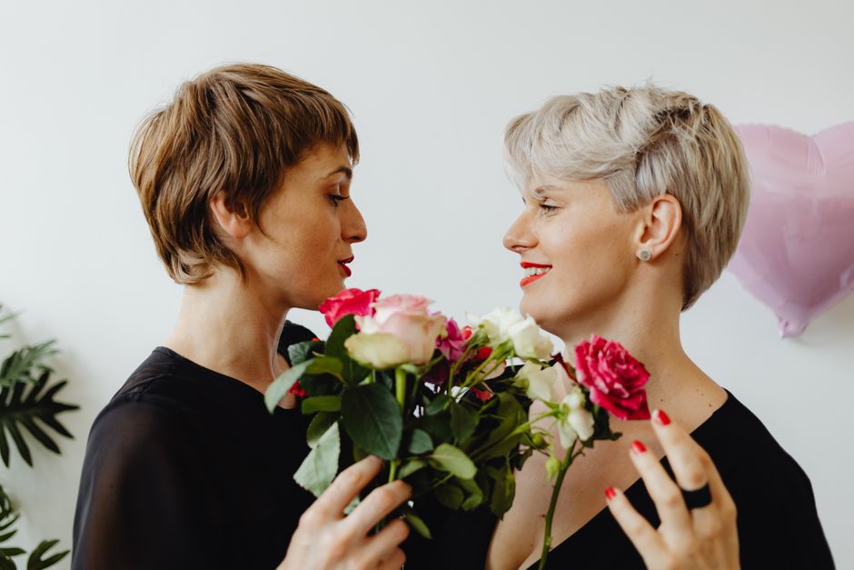 A close-up of two women with short hair, one with light brown and the other with silver hair, standing face-to-face in a bright, minimal indoor setting. They are exchanging a bouquet of fresh flowers,