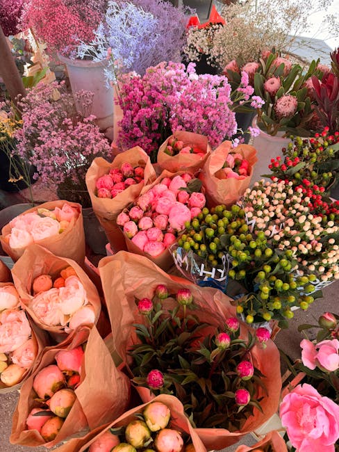 A vibrant display of freshly cut flowers and blooms arranged in various styles, featuring pink and peach peonies, clusters of pink and red berries, and sprigs of baby's breath and lavender. The flower