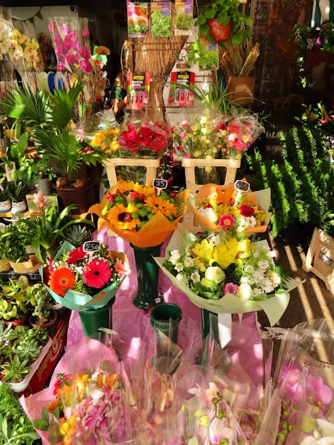 A vibrant display of fresh floral arrangements and bouquets arranged on a pink cloth-covered table in a flower shop. The bouquets feature bright pink gerbera daisies, yellow sunflowers, white roses, a